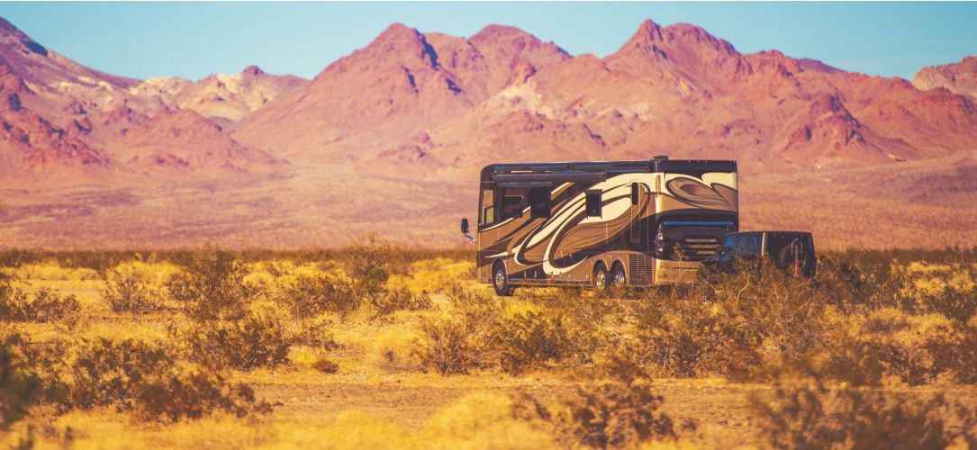Large RV towing a vehicle behind it in a Utah landscape with dry grass and red rocky mountains in the distance